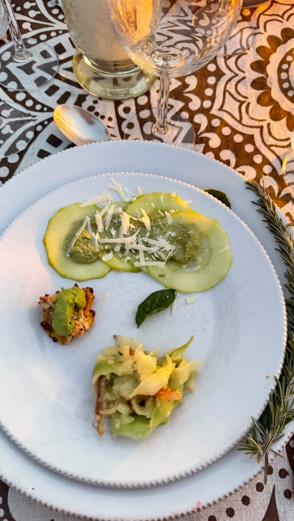 A plate with a green sauce and cucumber slices topped with cheese, accompanied by a small serving of seasoned vegetables and a bite-sized piece of fried food, with a decorative tablecloth and glasses in the background.