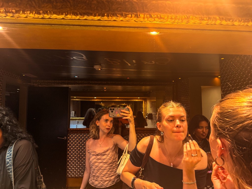 A group of women applying makeup in a stylish bathroom with mirrors and artistic decor, showcasing their reflections.
