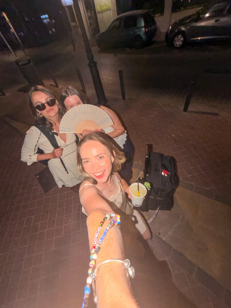 Three women posing for a selfie on a street at night, one holding a fan and another holding a drink, with parked cars in the background.