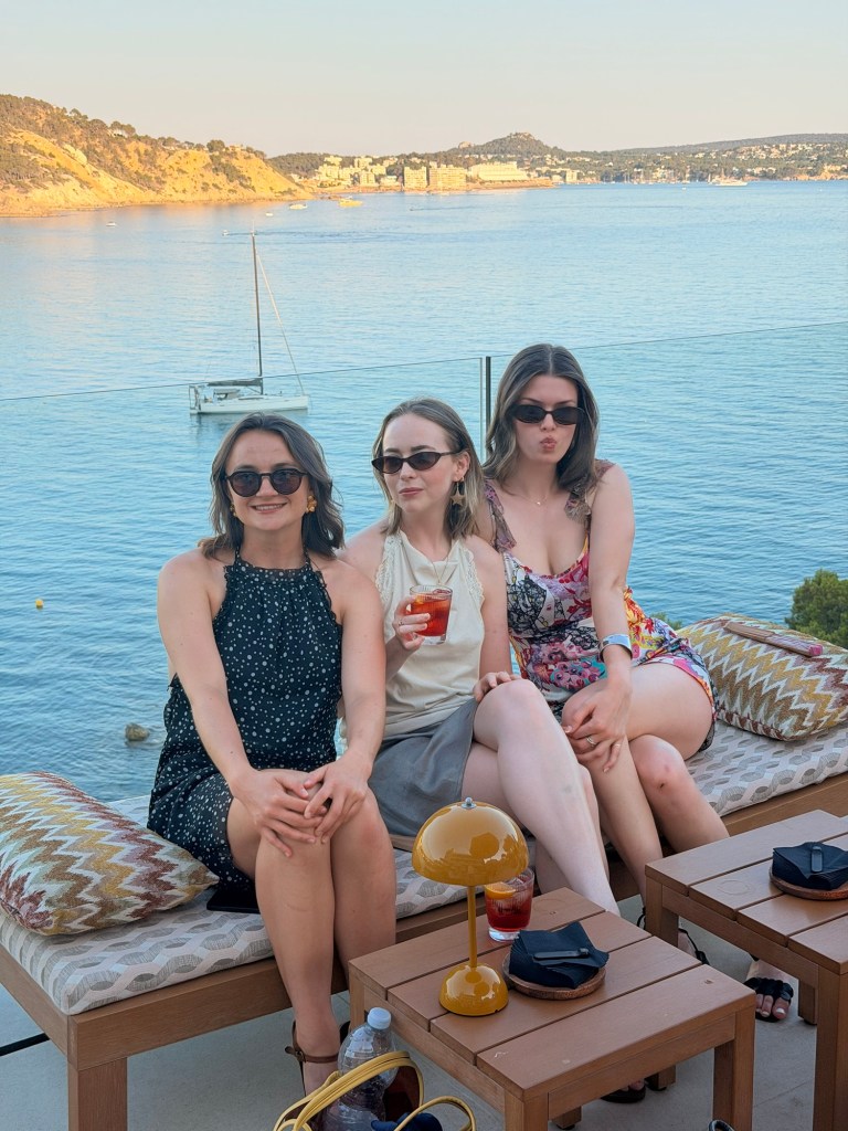 Three women sitting on a couch with a scenic view of the water and hills in the background. They are wearing sunglasses and enjoying drinks.