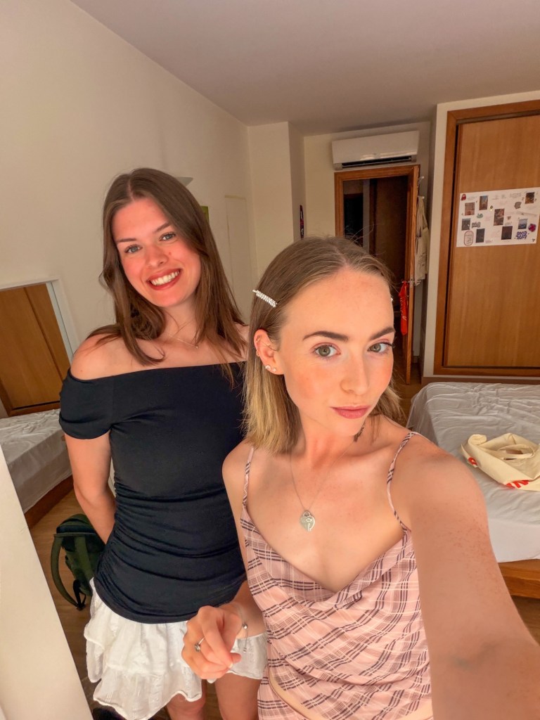 Two young women posing together in a room, with one wearing a black off-shoulder top and the other in a pink patterned top. The background features a bed and wooden wardrobe.