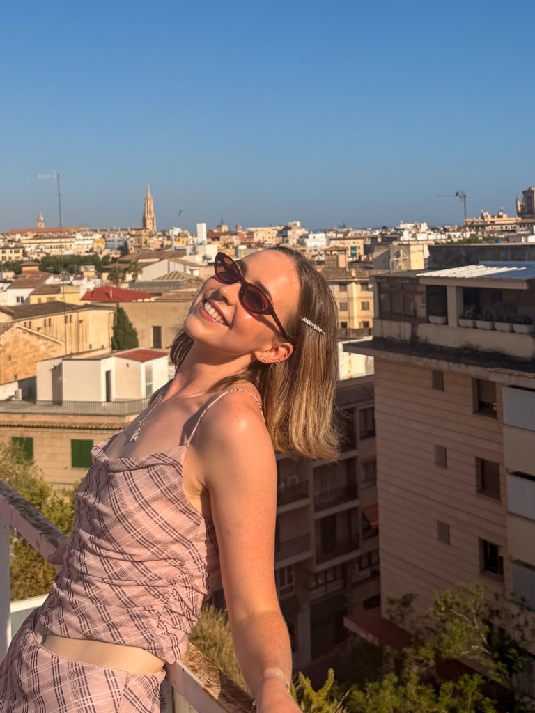 A woman with short hair, wearing sunglasses and a patterned outfit, smiles joyfully while leaning on a balcony surrounded by a cityscape under a clear blue sky.