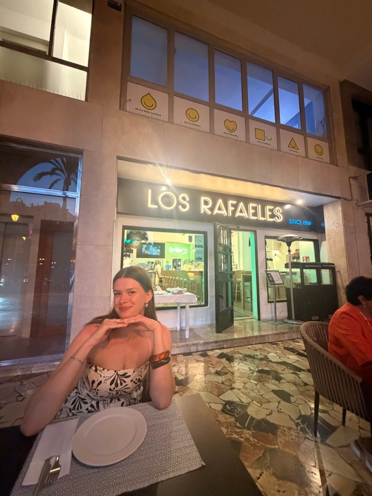 A young woman sitting at a table in front of a restaurant called 'Los Rafaeles', with her hands under her chin and smiling. The restaurant is brightly lit and features a modern design.