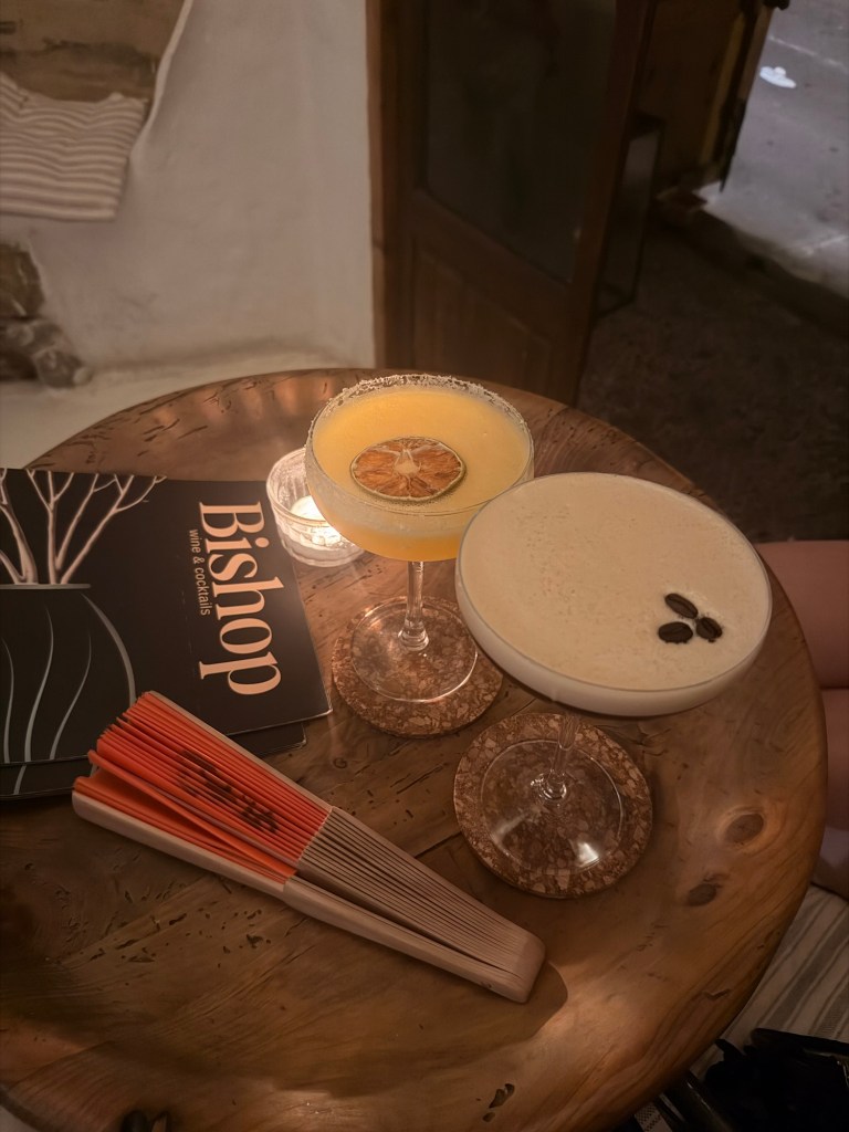 A wooden table featuring two cocktails: a yellow drink with a dried orange slice and a creamy drink topped with coffee beans, alongside a cocktail menu and a set of decorative chopsticks.