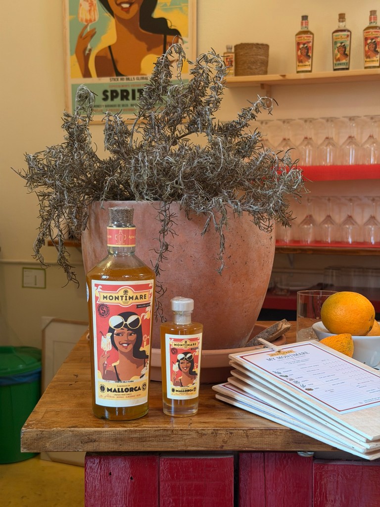 A wooden table displaying two bottles of Montimare, one large and one small, alongside a menu, fresh oranges, and a large terracotta pot filled with dried herbs. In the background, a vibrant poster depicting a woman with sunglasses and a relaxed pose adds colour to the scene.