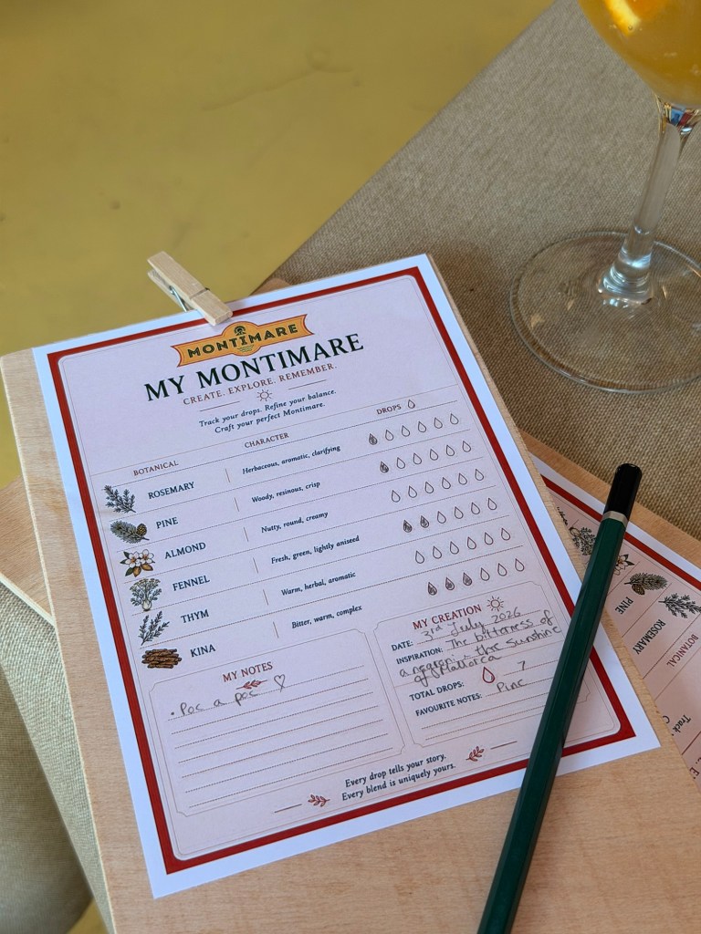A close-up of a Montimare crafting sheet featuring sections for selecting botanical flavours like rosemary, pine, almond, fennel, thyme, and kina. The sheet includes space for personal notes and a description of a unique creation, alongside a green pen and a drink glass in the background.