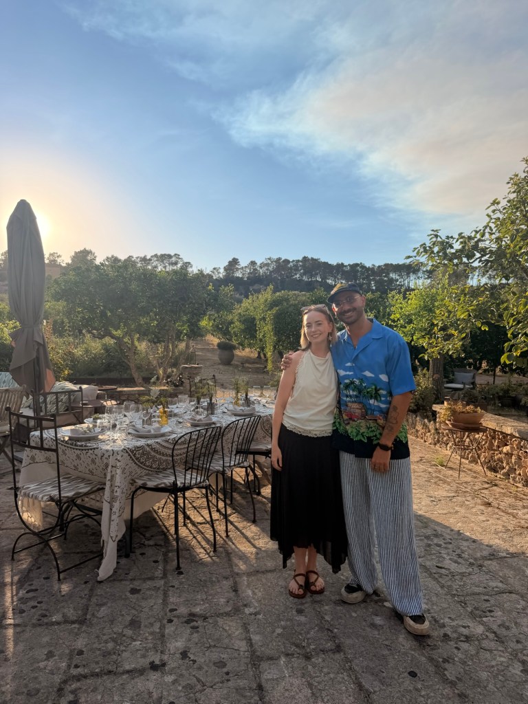 A woman in a white top and black skirt and a man in a colourful shirt and striped trousers pose together outdoors, with a table set for a meal in the background, surrounded by greenery and under a clear sky.