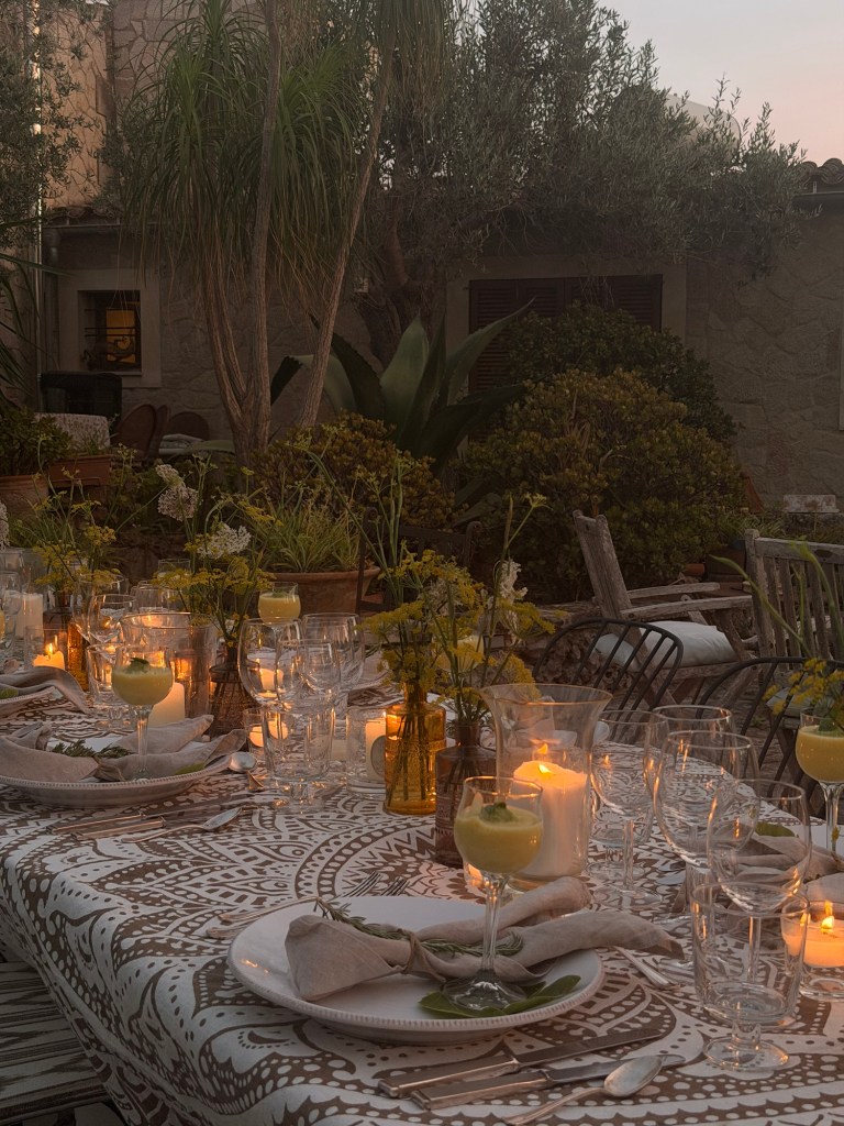 A beautifully set outdoor dining table with glasses, plates, and stylish cutlery. Yellow drinks are served in elegant glasses, surrounded by small vases of flowers and glowing candles, with lush greenery in the background.