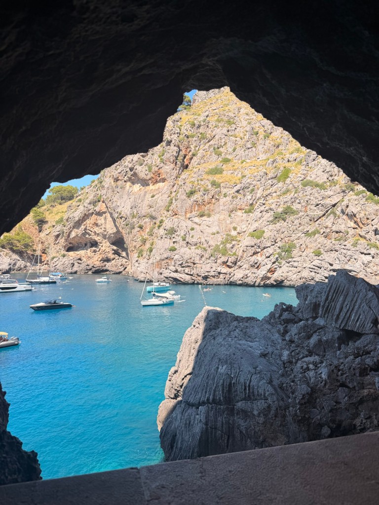 View of a serene bay with moored boats, framed by rocky cliffs and vibrant blue water, seen through a natural archway.