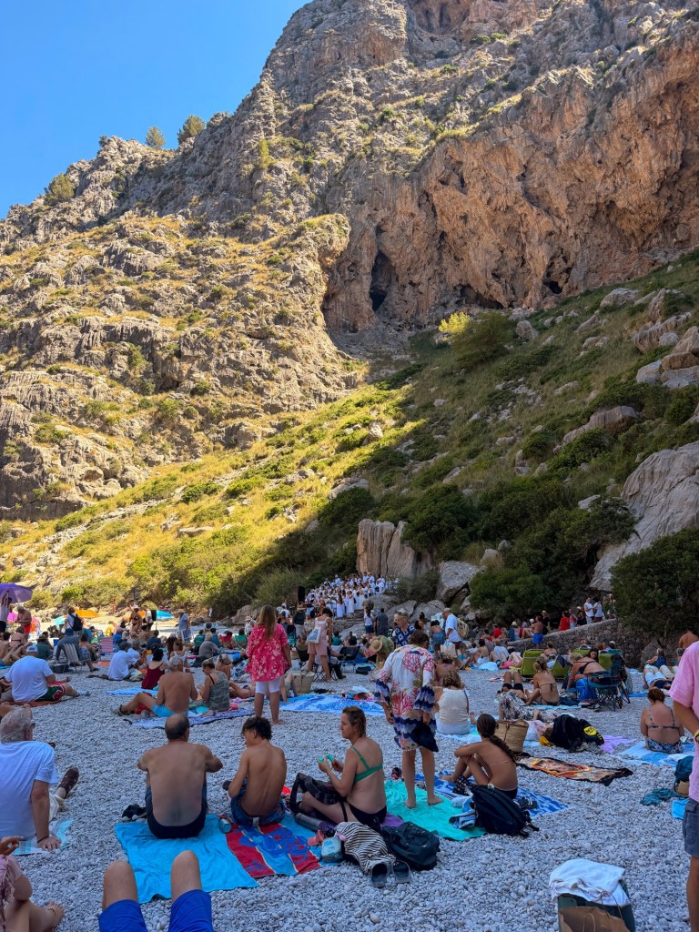 A crowded beach scene with sunbathers on towels, surrounded by rocky cliffs and greenery under a clear blue sky.