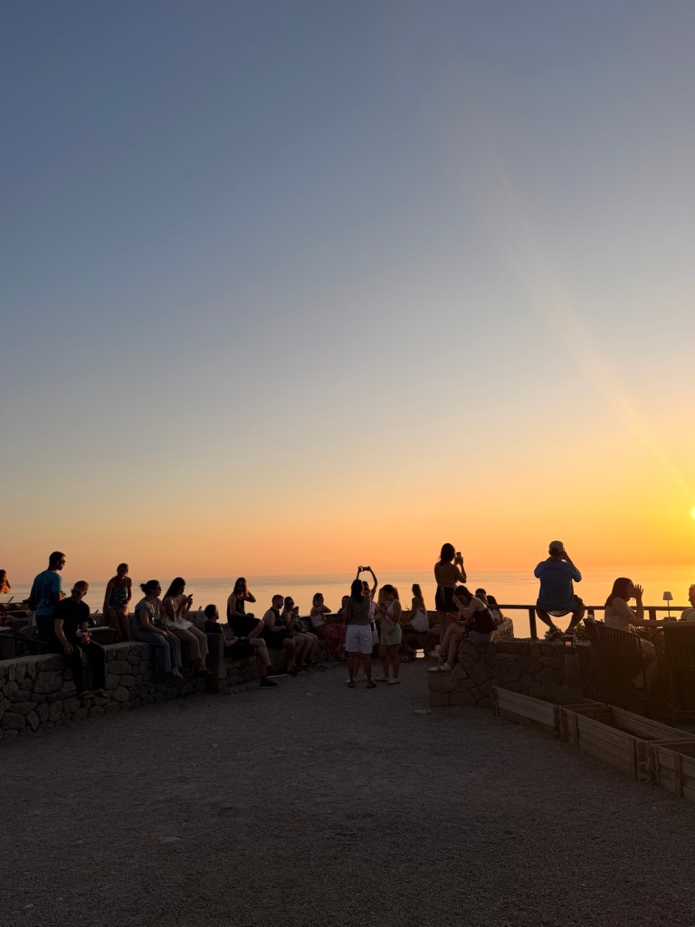 People gathered on a rocky shoreline at sunset, enjoying the view and taking photos.