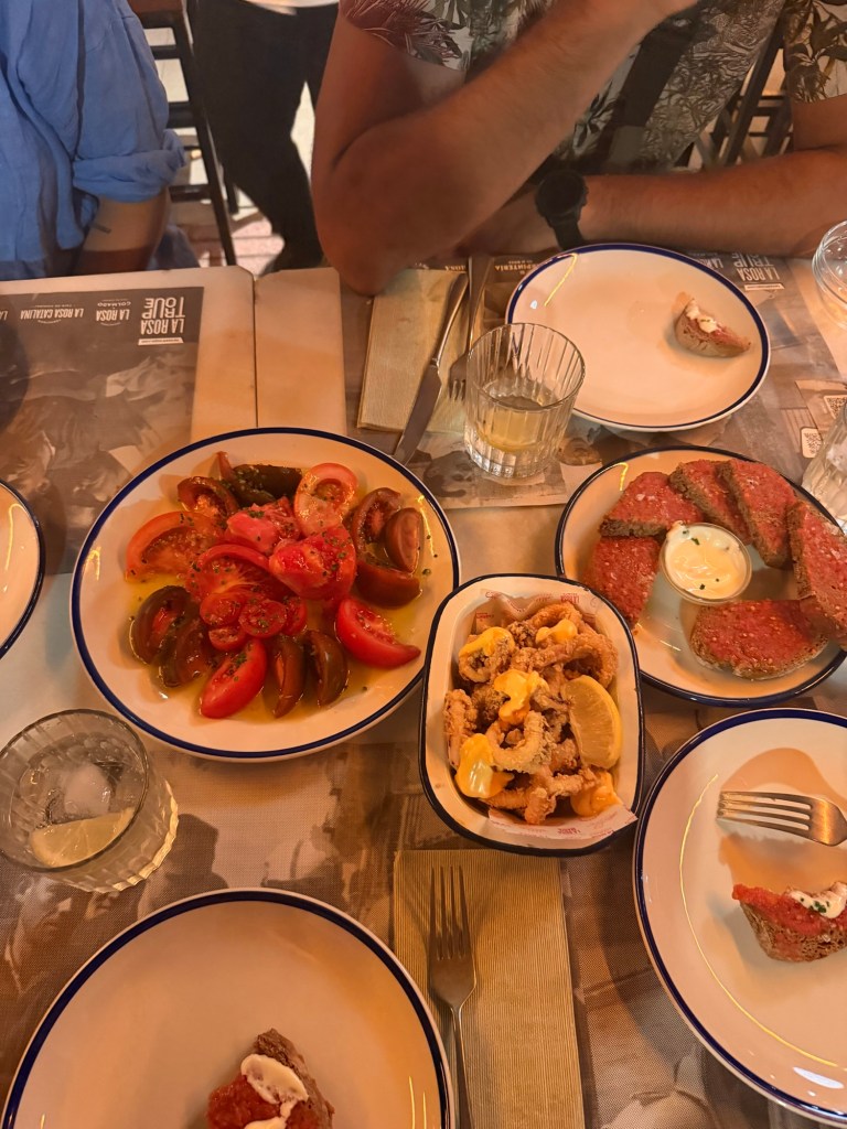 A spread of various dishes on a dining table, featuring a plate of sliced tomatoes drizzled with olive oil, a serving of fried calamari topped with sauce and lemon, and several pieces of bread with toppings. Empty plates and glasses are also visible.